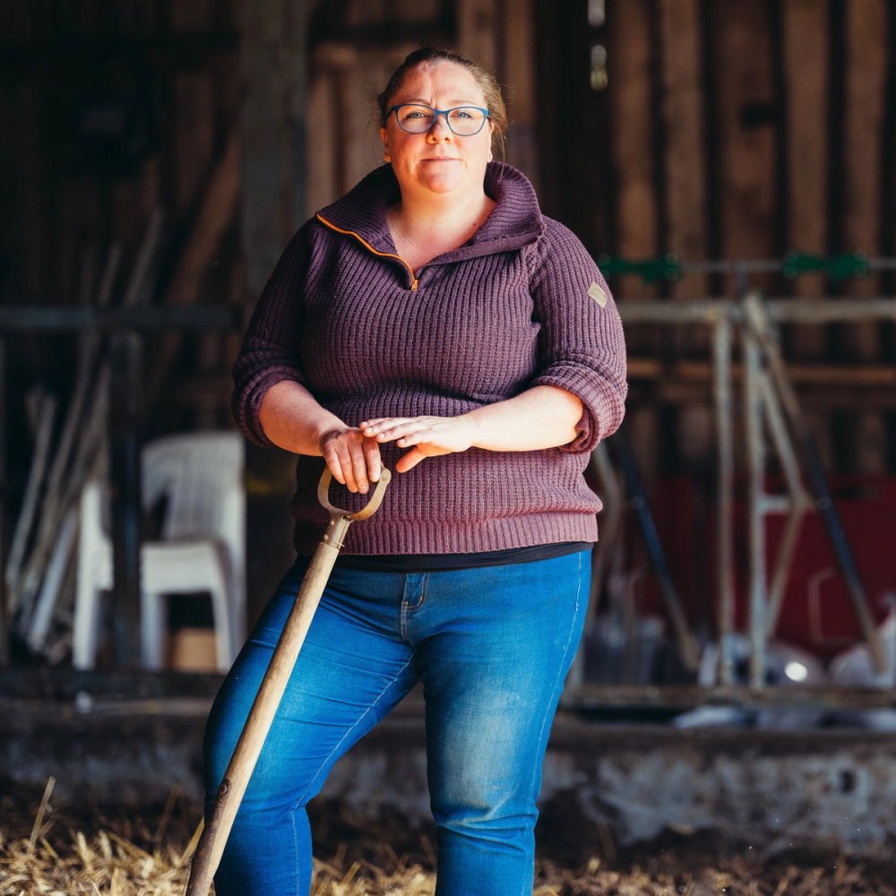 Person med bløt genser og jeans holder en spade i en låve eller stall med treverk i bakgrunnen.
