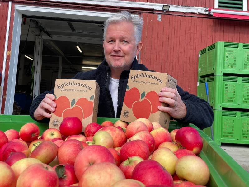 Person som selger epler foran en butikk med eplekartonger og frukt på bordet, med synlige haglskader på epleblomster i bildet.