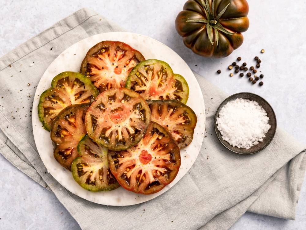 Skiver av tomat på en hvit tallerken, med salt og pepper ved siden av, på et lyst bord.