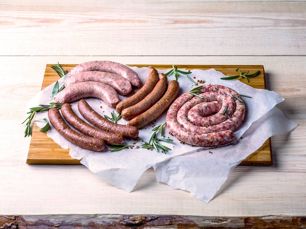Ulike typer pølser, inkludert rå pølser, lagt på et skjærebrett med noe grønt urtekrydder, klar til tilberedning.