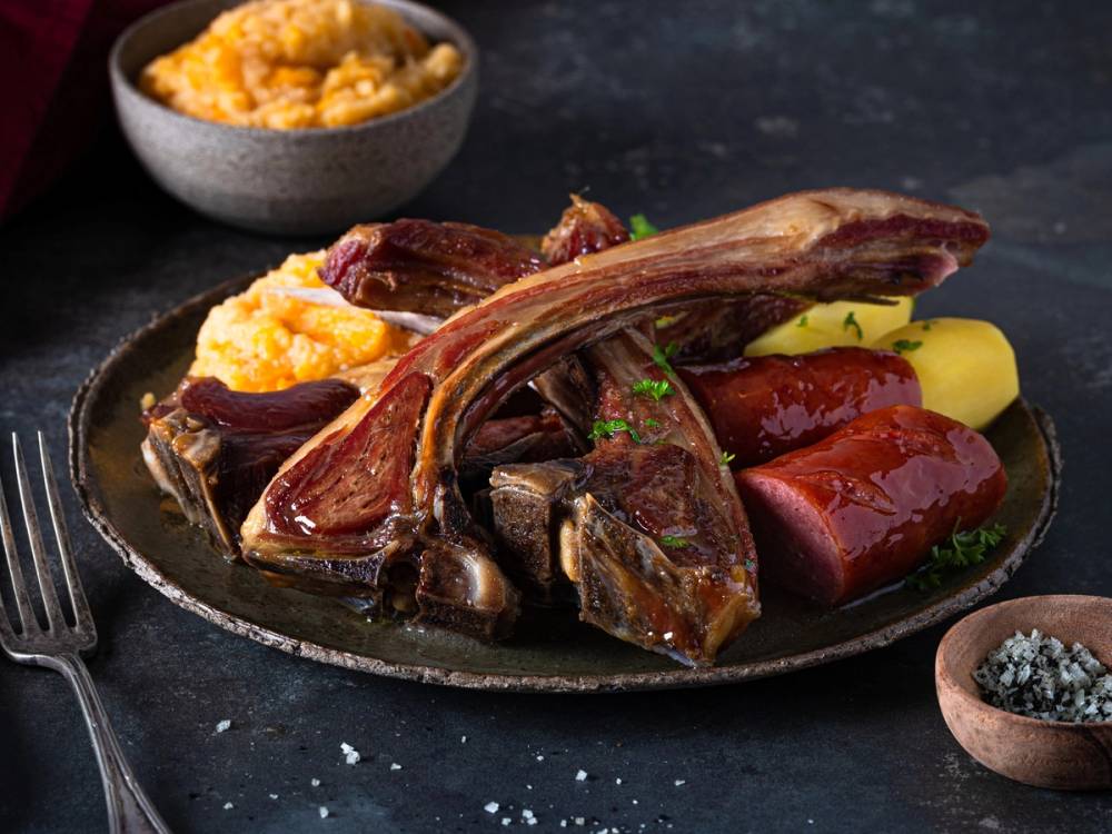 Pinnekjøtt med kålrotstappe og poteter på et svart fat.