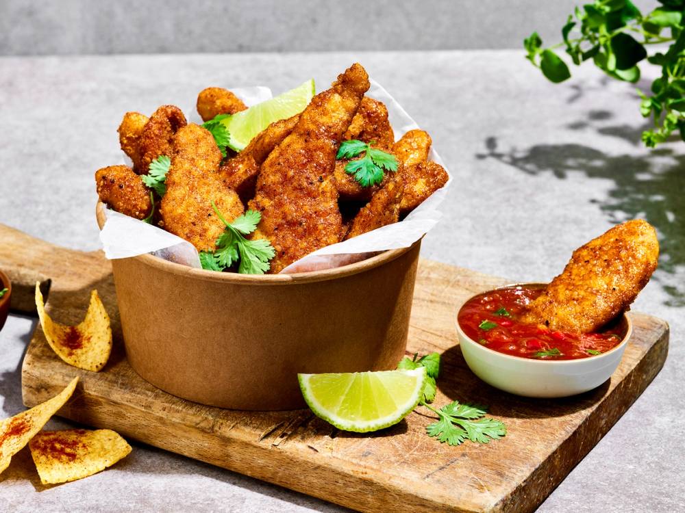 Krispy kyllingstikker med salsa og lime på et trebord, omgitt av potetgull og urter.