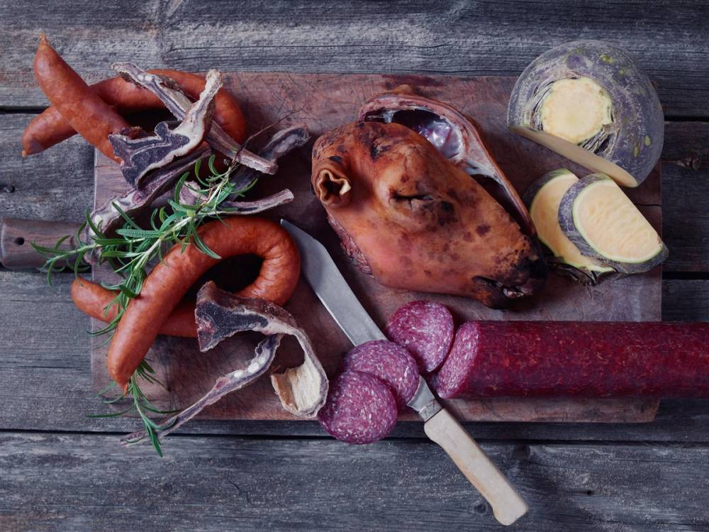 Eldhusprodukter som pølser, spekepølse, ost og en skive brød på et trebenet bord.