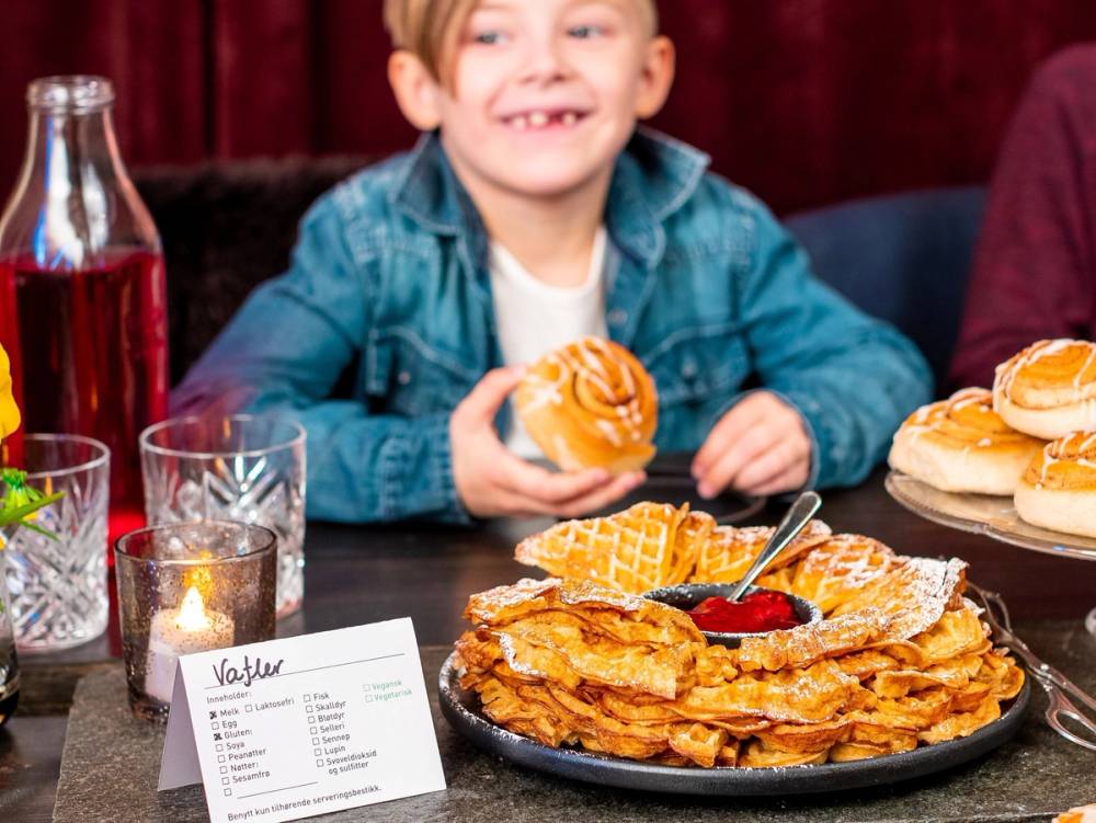 Barn som spiser vafler og har en tallerken med flere pannekaker på bordet, omgitt av drikke og lys.