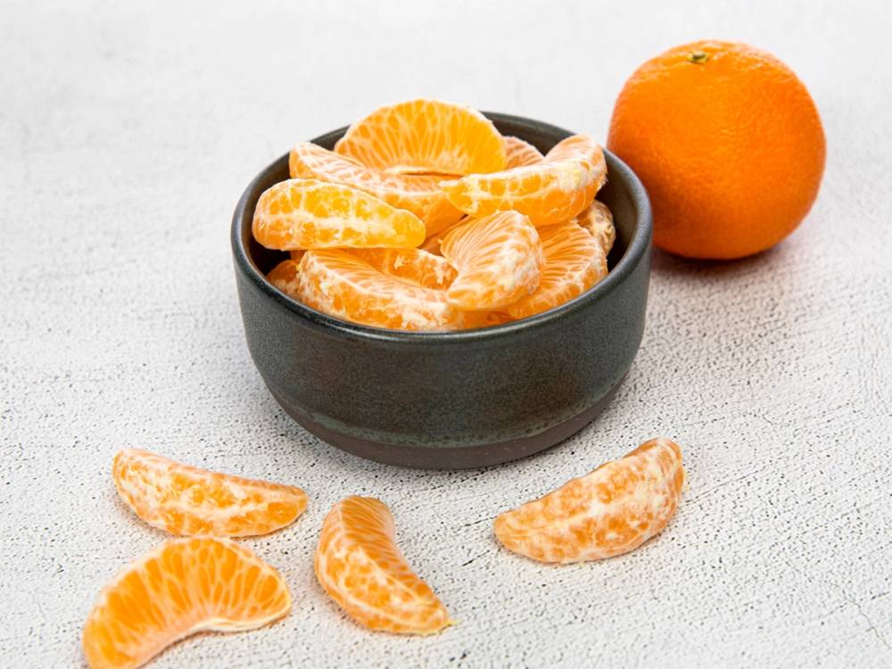 En svart skål med skrelte mandariner og en hel appelsin ved siden av på et hvitt underlag.