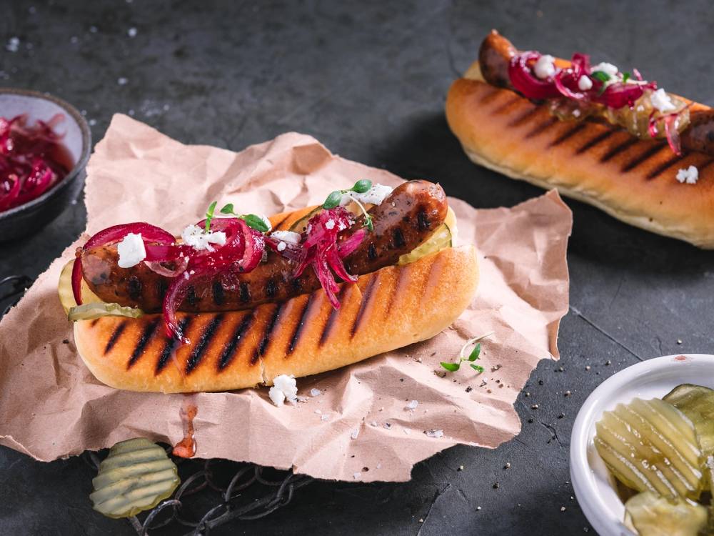 Lammepølse med syltet rødløk i en grillett, servert på papir med tilbehør i små boller rundt omkring.