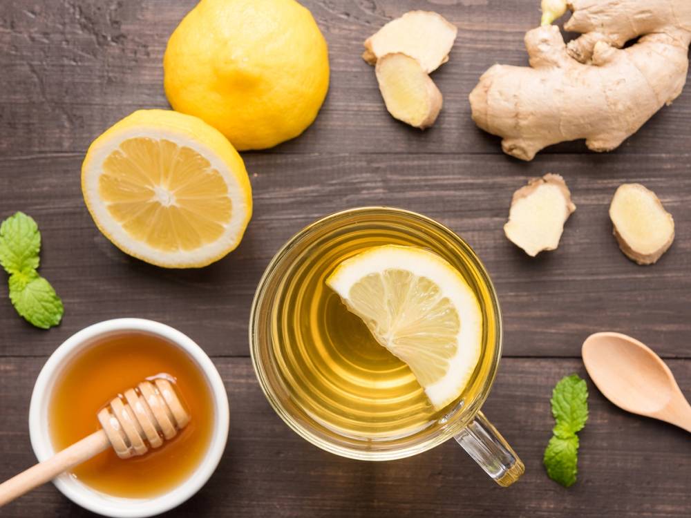 Sitron, ingefær og honning i en kopp, med fersk sitron og ingefær på en treplate, samt noen mynteblader og en skje, på et mørkt trebord.