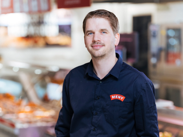 Morten, driftsjef, står i en matbutikk med en Meny-navneskilt på skjorta, omgitt av ferske varer og hyller.