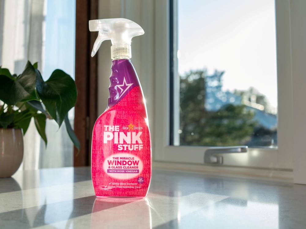 En rosa sprayflaske med etiketten 'The Pink Stuff' står på et bord foran et vindu.