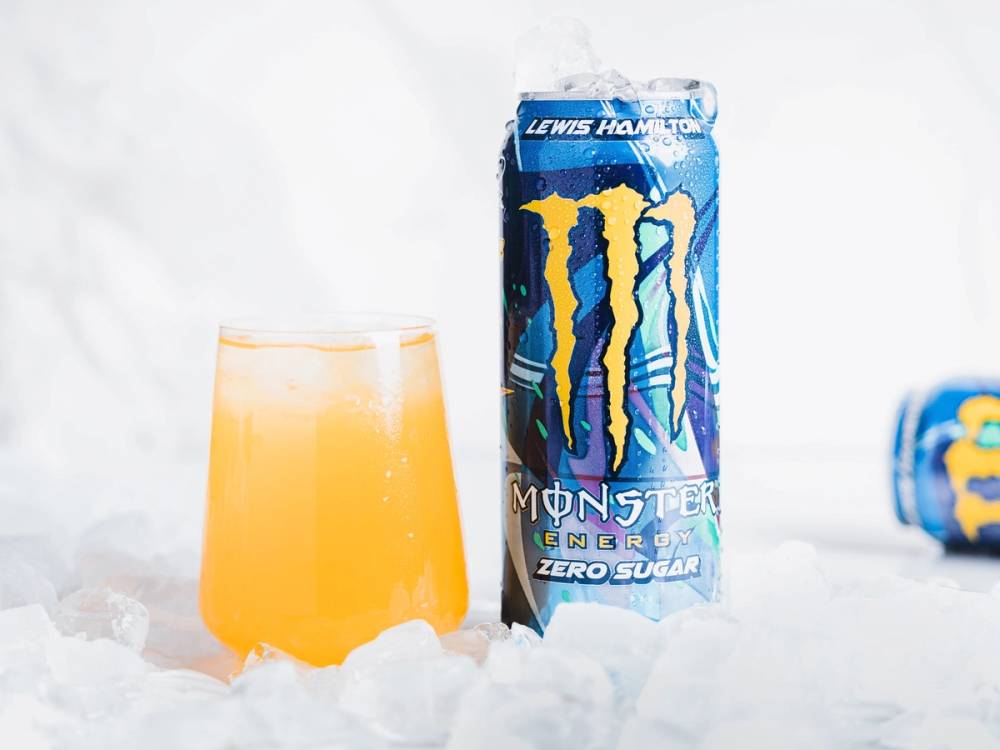 En energidrikksboks med Monster-logo og en glass med oransje drikke ved siden av, plassert på snøaktig underlag.