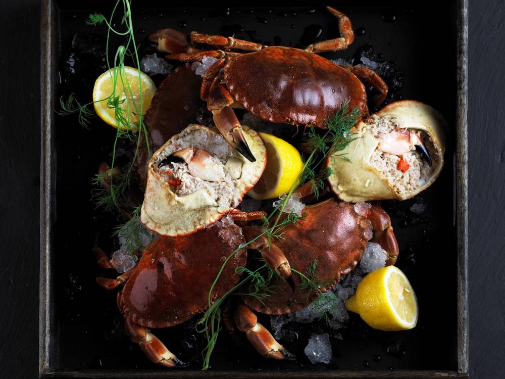 Slik-tilbereder-du-krabbe-krabbelag.jpg viser ferske krabber med sitronbåter og urter på en svart overflate.