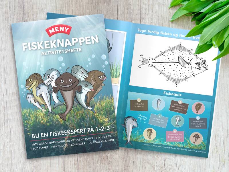 Et aktivitetshefte med fisketema, med bilder av fisk og aktiviteter, ligger på et bord ved siden av grønne blader.