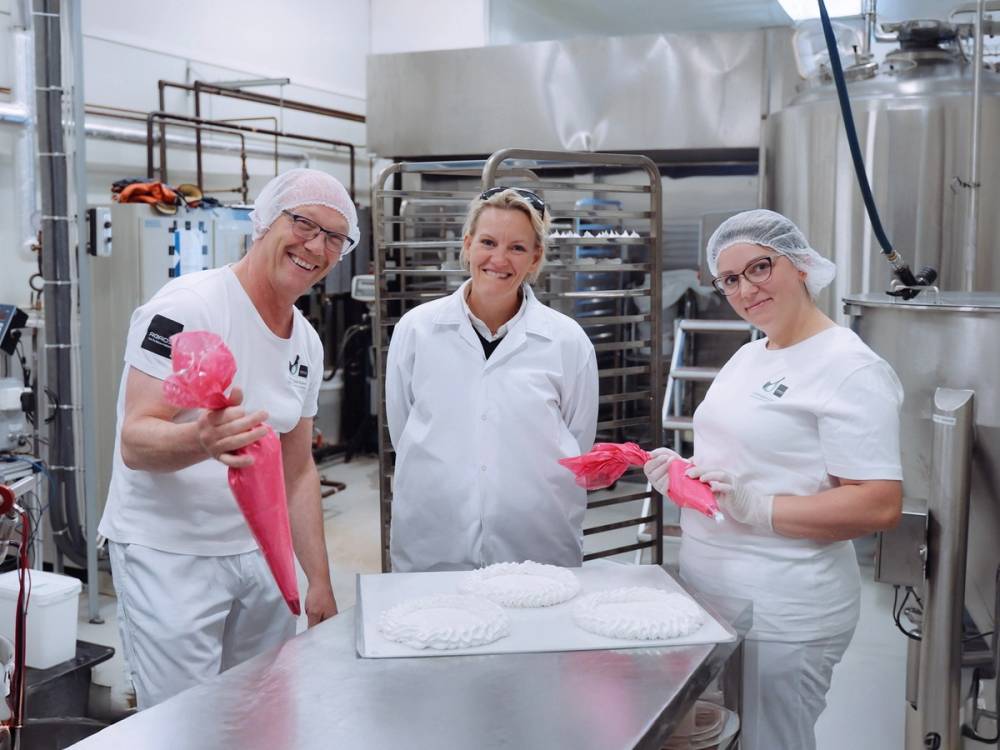 Tre personer i hvite arbeidsklær og rosa hansker står rundt et bord i et kjøkken eller bakeri, med metallutstyr og hyller i bakgrunnen.