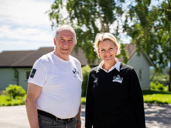 To personer står ute foran en gård med grønne trær og hus i bakgrunnen, begge kledd i hvite og svarte klær med logoer.