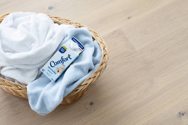 En kurv med hvite og lyseblå klær, inkludert et plagg med Comfort-merke, plassert på et lyst tregulv.