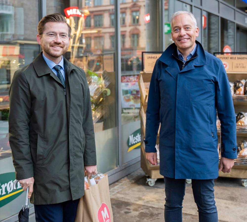 To personer står utenfor en butikk, en av dem holder en handlepose, og de er kledd i jakker.