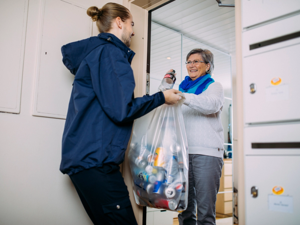 To personer utveksler en pose med tomflasker foran en dør, i en innendørs setting.