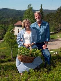 To personer står sammen i en grønn hage med en kurv med urter, omgitt av trær og fjell i bakgrunnen.