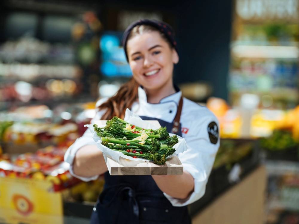 En person i butikkuniform som holder en tallerken med grønnsaker, sannsynligvis asparges, i en dagligvarebutikk.
