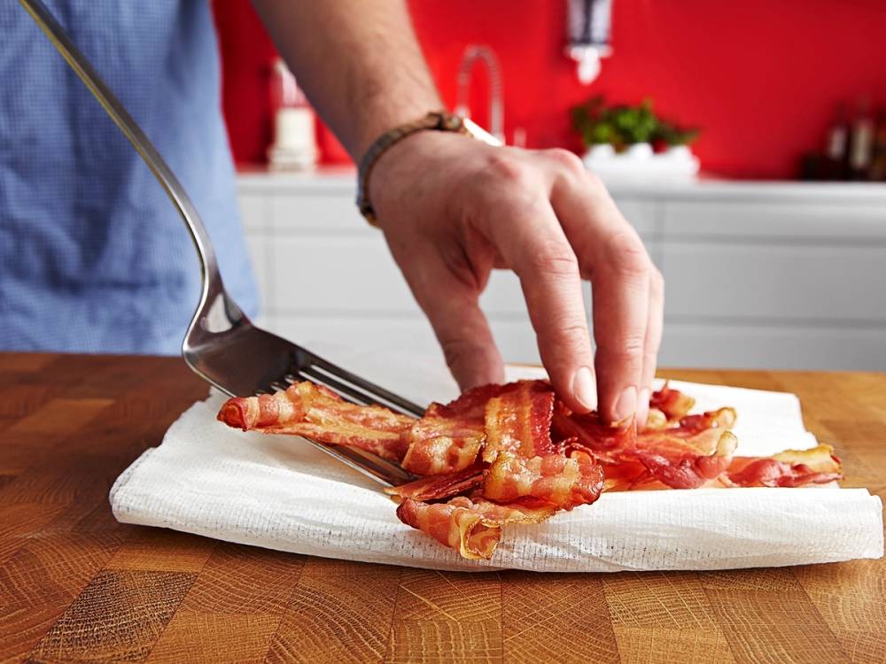 En person legger skiver av bacon på en hvit papirhåndkle på et kjøkkenbord.