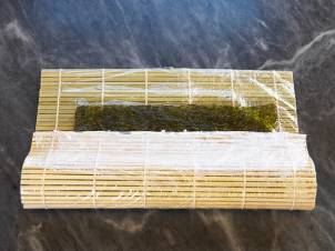 En rull med sushi-ingredienser, der en nori-ark er lagt på en bambusmatte klar for rulling.