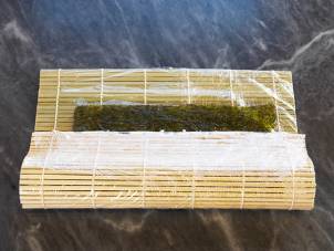 En rull med sushi-ingredienser, der en nori-ark er lagt på en bambusmatte klar for rulling.