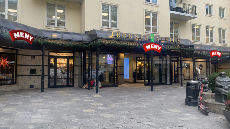 Fasaden til Frogner Atrium med MENY-butikk og inngangsparti, omgitt av en åpen plattform og noen sykler og søppelbøtter.