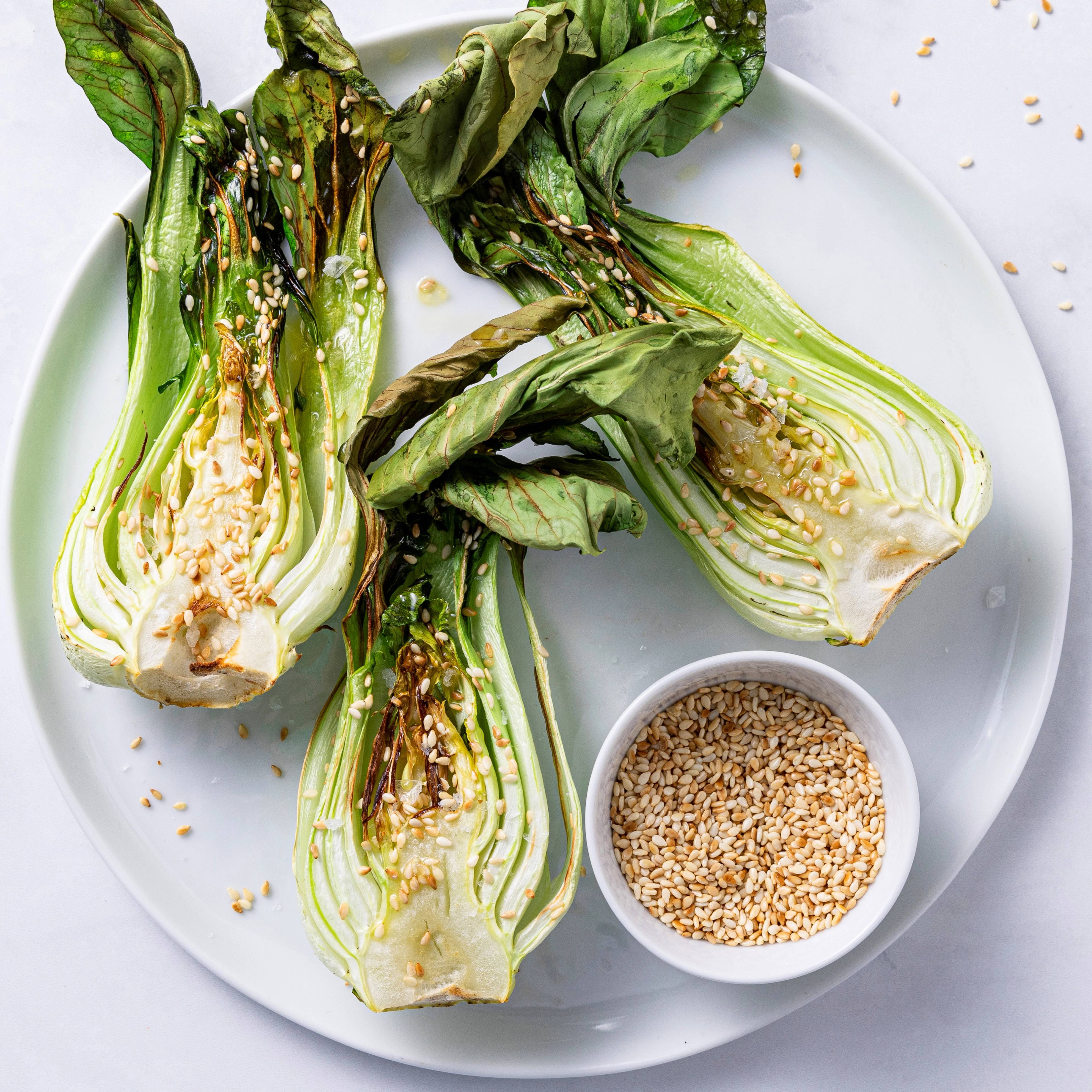 Pak choy med sesamolje og sesamfrø på en hvit tallerken, med en liten skål med sesamfrø ved siden av.