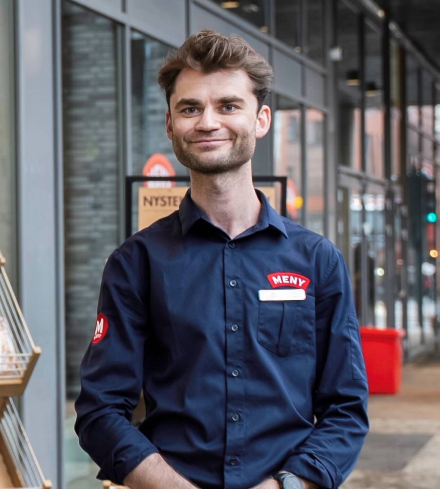 En ansatt i Meny-butik står inne i butikken med butikkuniform og navnplate, foran butikkens inngang.