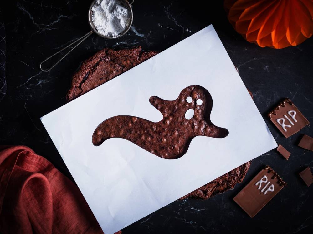 En sjokoladekake i form av en dinosaur på et hvitt ark, omgitt av Halloween-dekorasjoner som en liten gravstein og en oransje papirlue, med en måne i bakgrunnen.