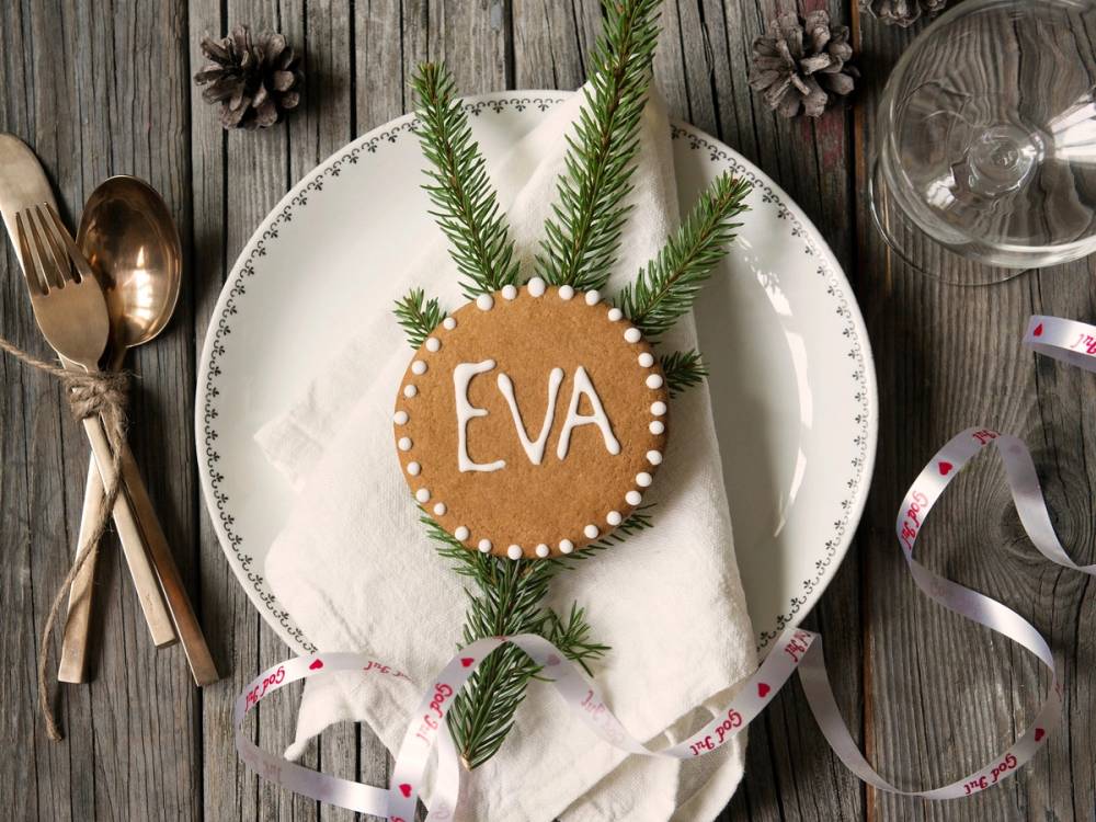 Juledekorasjon med et pepperkake med navnet Eva, pyntet med grønne granbar og små hvite prikker, plassert på en hvit tallerken med sølvbestikk og julepynt rundt.
