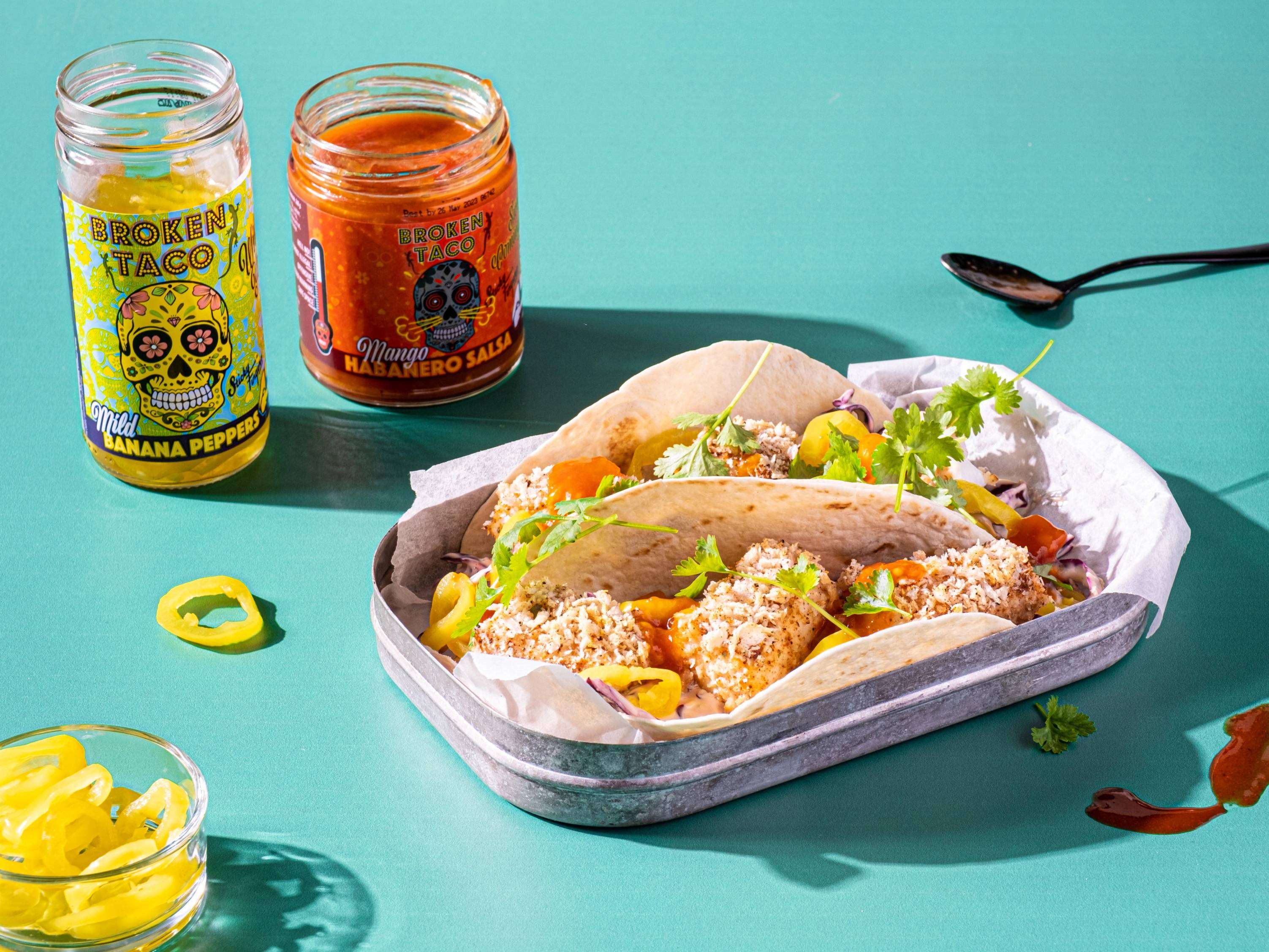 Fisketacos med grønnsaker og sitron på et fat, med to glass med salsa og en skje ved siden av, på en turkis bakgrunn.