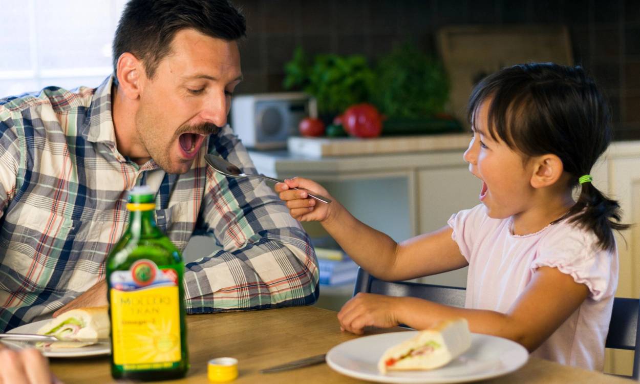 En voksen og et barn sitter ved et kjøkkenbord, og barnet gir voksen en tablett eller medisin, med en flaske på bordet.