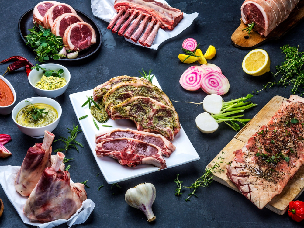 Ulike typer av kjøtt, blant annet lam, ribbe og pølser, er lagt ut på en mørk overflate med urter, grønnsaker og sauser rundt.
