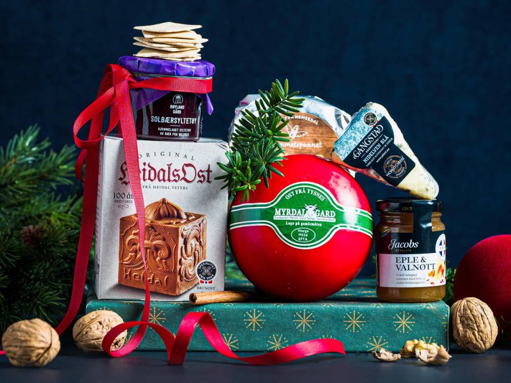 Utsikt av ulike matgaver, inkludert ost, sjokolade og syltetøy, dekorert med granbar og nøtter, på en julepyntet bakgrunn.