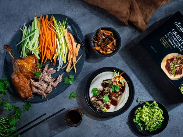 En tallerken med kyllingvinger, grønnsaker og en liten skål med saus, omgitt av andre matretter og en pakke med mat på et mørkt bord.