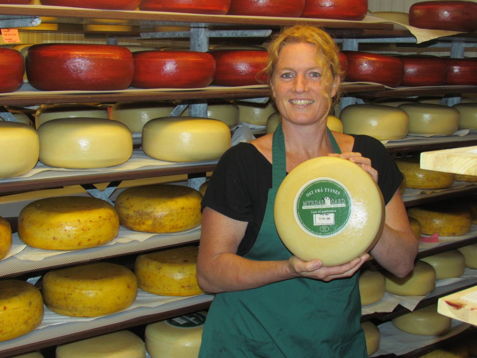 En person holder et stort ostestykke foran en vegg med mange ostekuler i ulike farger og størrelser på en ostebutikk.