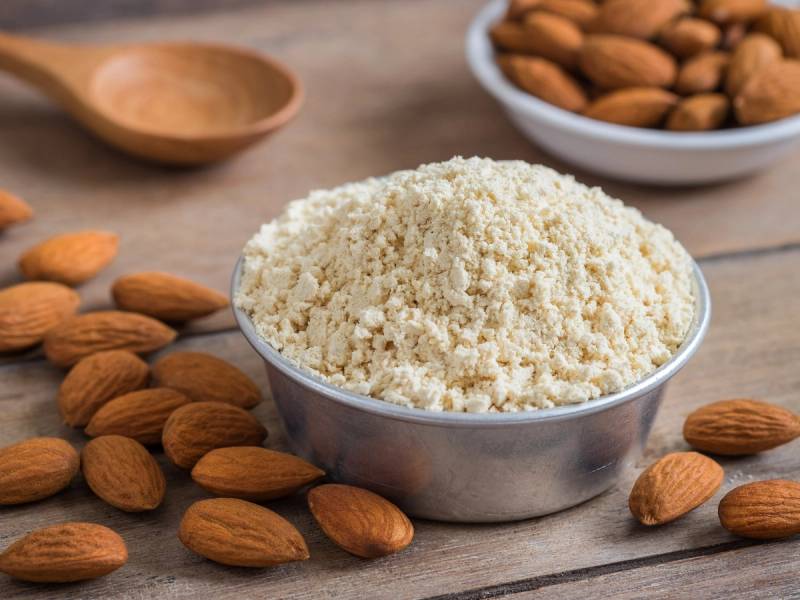 Håndfull mandelmel i en bolle med flere hele mandler rundt på et trestykke bord