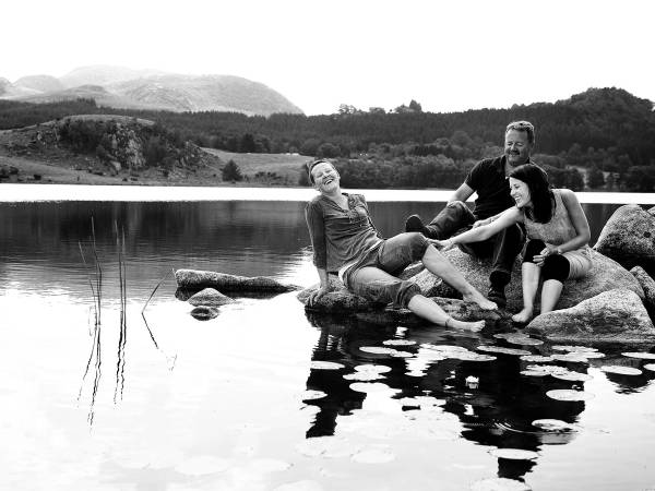 Tre personer sitter på store steiner i vannet ved en innsjø, omgitt av natur med fjell i bakgrunnen.