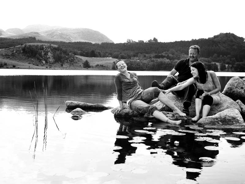 Tre personer sitter på store steiner i vannet ved en innsjø, omgitt av natur med fjell i bakgrunnen.