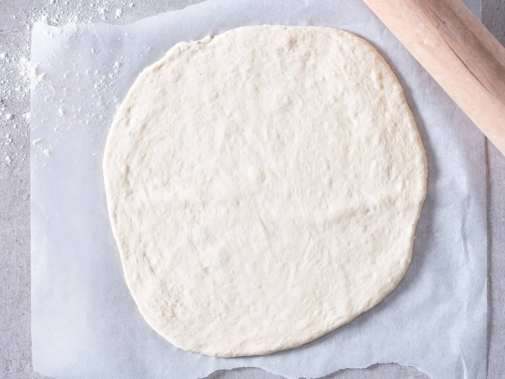 Rund pizzadeig som er utkjevlet og ligger på bakepapir klar for topping.