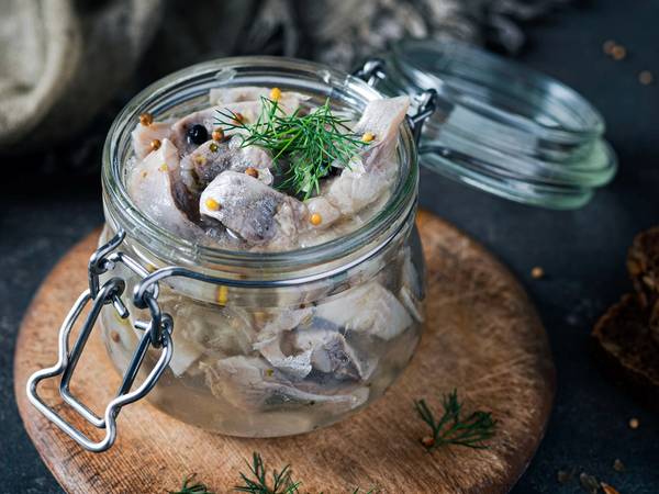 Sursild i glasskrukke med dill på et rundt trebrikke, omgitt av brød og krydderurter.