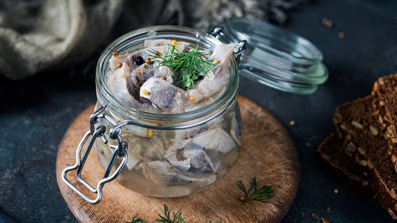 Sursild i glasskrukke med dill på et rundt trebrikke, omgitt av brød og krydderurter.