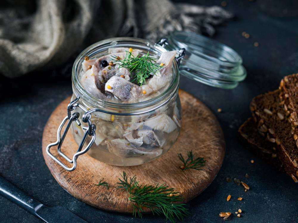 Sursild i glasskrukke med dill på et rundt trebrikke, omgitt av brød og krydderurter.