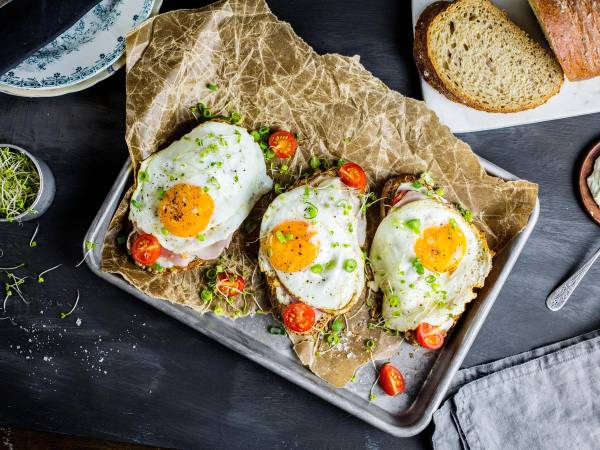 Tre stekte egg med cherrytomater og gressløk på et bakepapir på en metallplate, med brød i bakgrunnen.