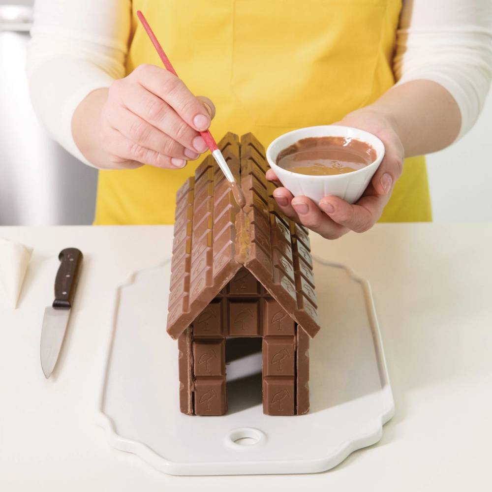 Person som dekorerer et sjokoladehus med en pensel og har en skål med sjokolade ved siden av, på et hvitt bord.