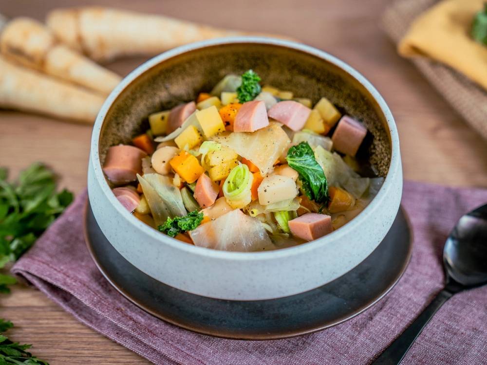 Grønnsakssuppe med pølse i en skål på et bord med en skje ved siden av.