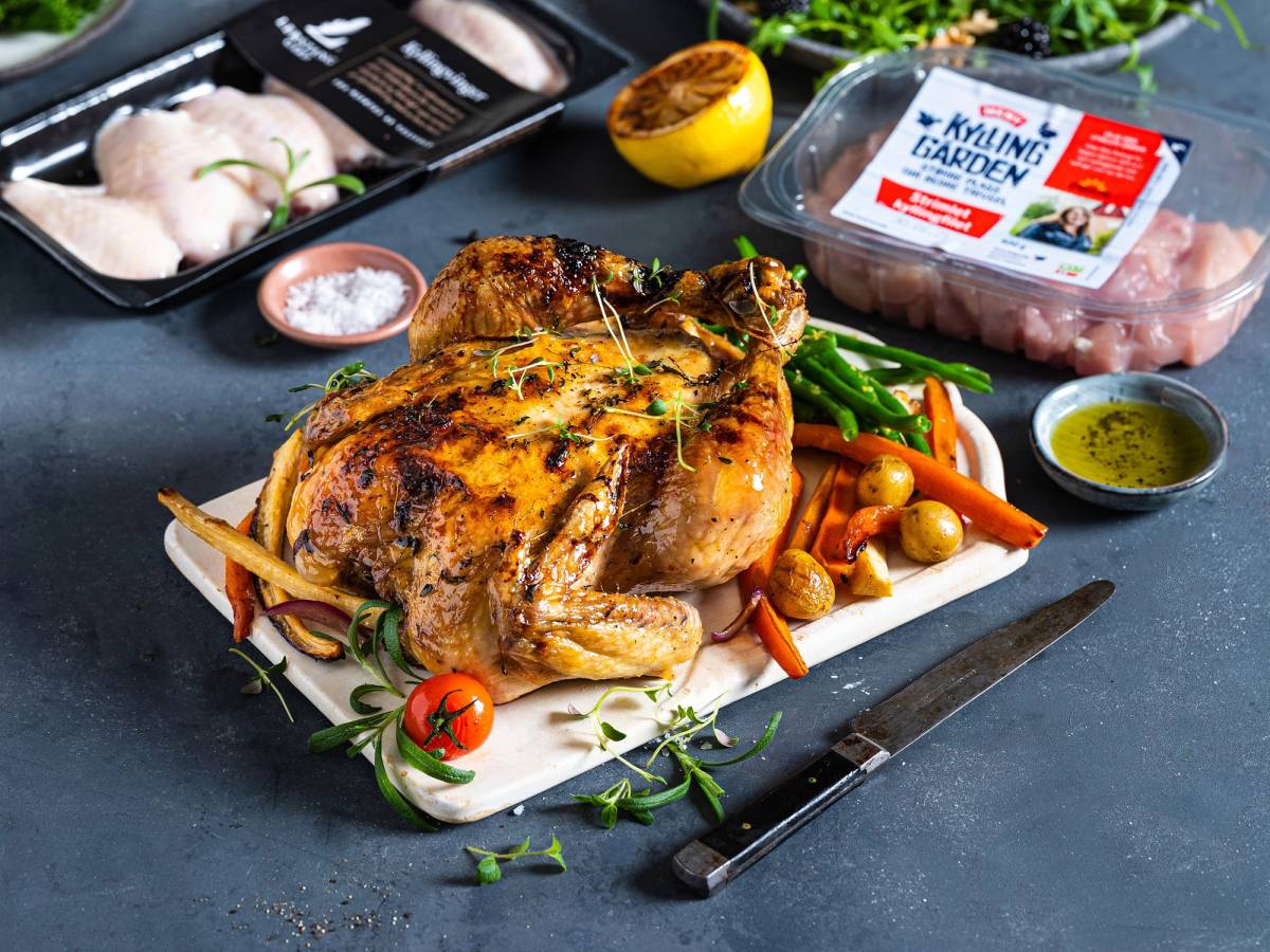 Helstekt kylling med grønnsaker og urter på et skjærebrett, omgitt av ingredienser og kjøkkenutstyr.