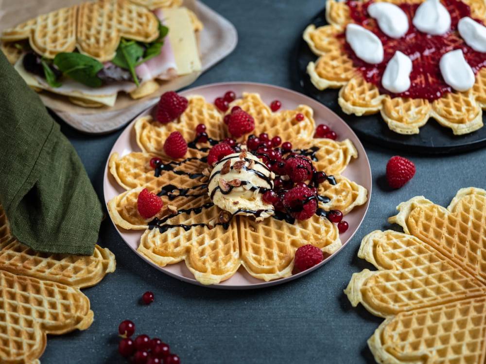 Vafler med syltetøy og is på en tallerken omgitt av flere ujevne vafler på en mørk bakgrunn.
