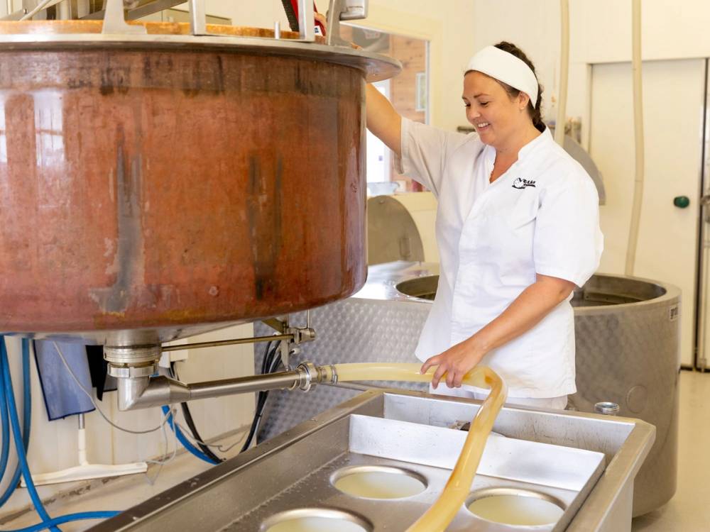 En person i hvit kokkeforkledning står ved en arbeidsbenk med en stor gryte eller kjele, og håndterer en lang skje eller redskap i et kjøkken eller matlagingsmiljø.